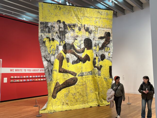 A large floor-to-ceiling textile hangs from the ceiling of an art gallery. The textile is yellow, grey, and brown and depicts individuals styling others' hair.