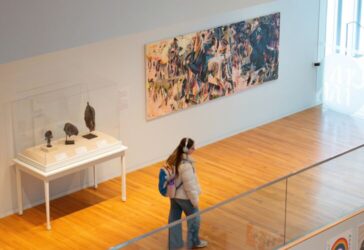 This image shows a modern art gallery with wooden floors and glass railings. Several artworks are displayed on the walls, including a large colorful abstract painting and smaller framed pieces. Bright natural light streams in from a window on the right.