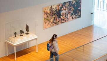 This image shows a modern art gallery with wooden floors and glass railings. Several artworks are displayed on the walls, including a large colorful abstract painting and smaller framed pieces. Bright natural light streams in from a window on the right.