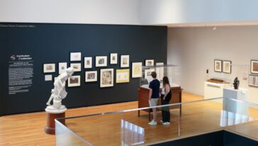 Two individuals viewing various artworks in a gallery room, including framed pieces on the wall and a sculpture displayed on a pedestal.