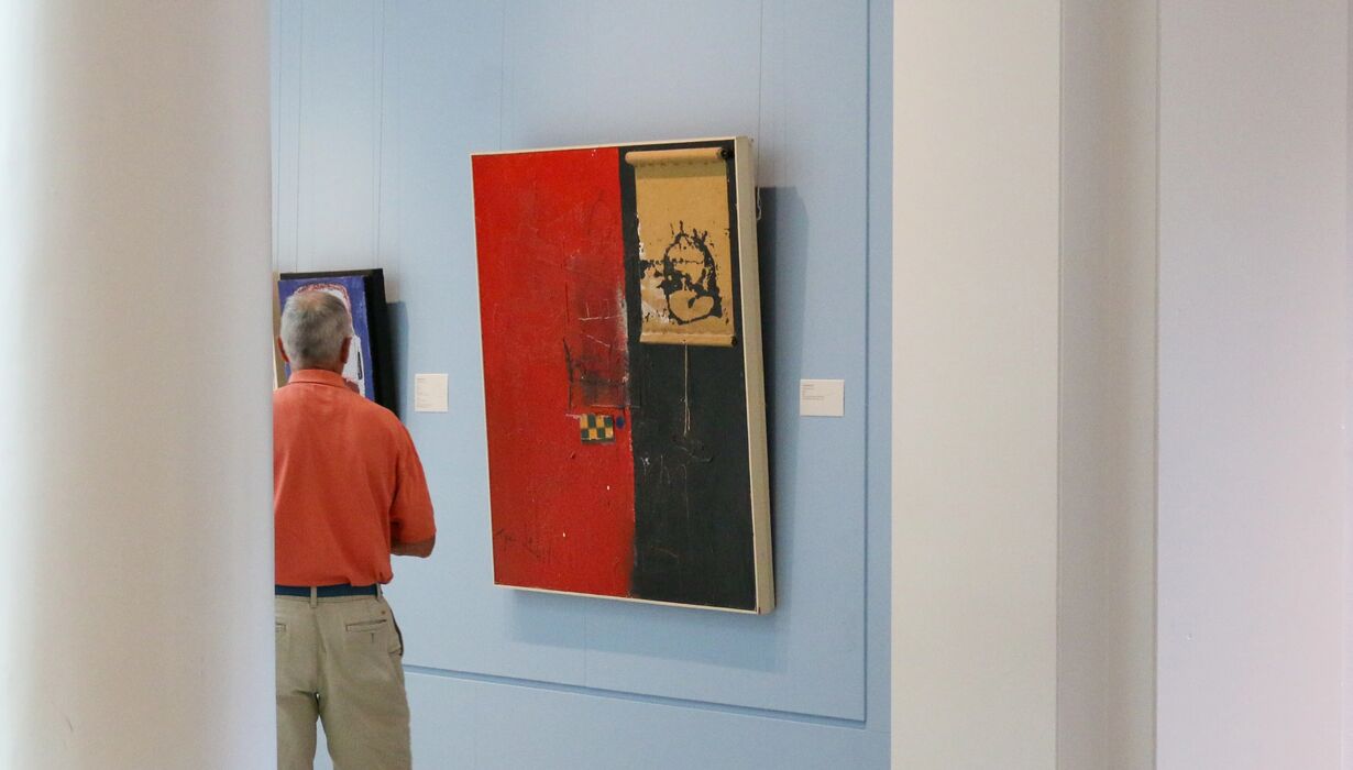 Person viewing abstract art pieces on display in a gallery, with one artwork prominently featuring red and black colors.