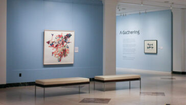 Art gallery interior showing an exhibition space with light blue walls and tiled floors. Two benches are centrally placed on the floor. To the left, a large colorful abstract painting is displayed, featuring vibrant reds and oranges with scattered elements of blue and pink. Adjacent to the right is a smaller framed artwork. A wall text titled "A Gathering" is visible on the right side.
