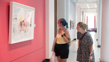 Two young female visitors examine a modern art piece that is hung against a bright pink wall.
