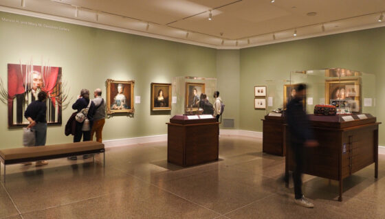 Groups of people stand and walk through a light green room with dark wooden vitrines and gilded framed paintings on the wall in the Unsettling Histories exhibition.