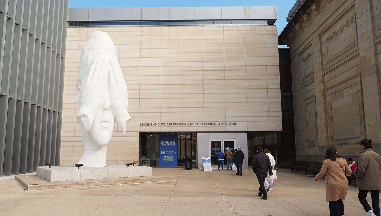 A line of people walk toward a museum entrance, and a large sculpture of a white head stands tall next to them.