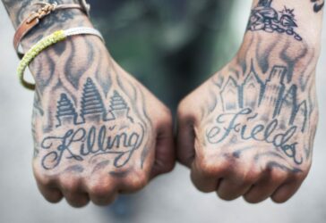 The tops of two fists which are covered in black and white tattoos. The left fist shows a drawing of the temple Ankgor Wat above the word "Killing". The right fist shows a drawing of the Philadelphia skyline above the word "Fields"