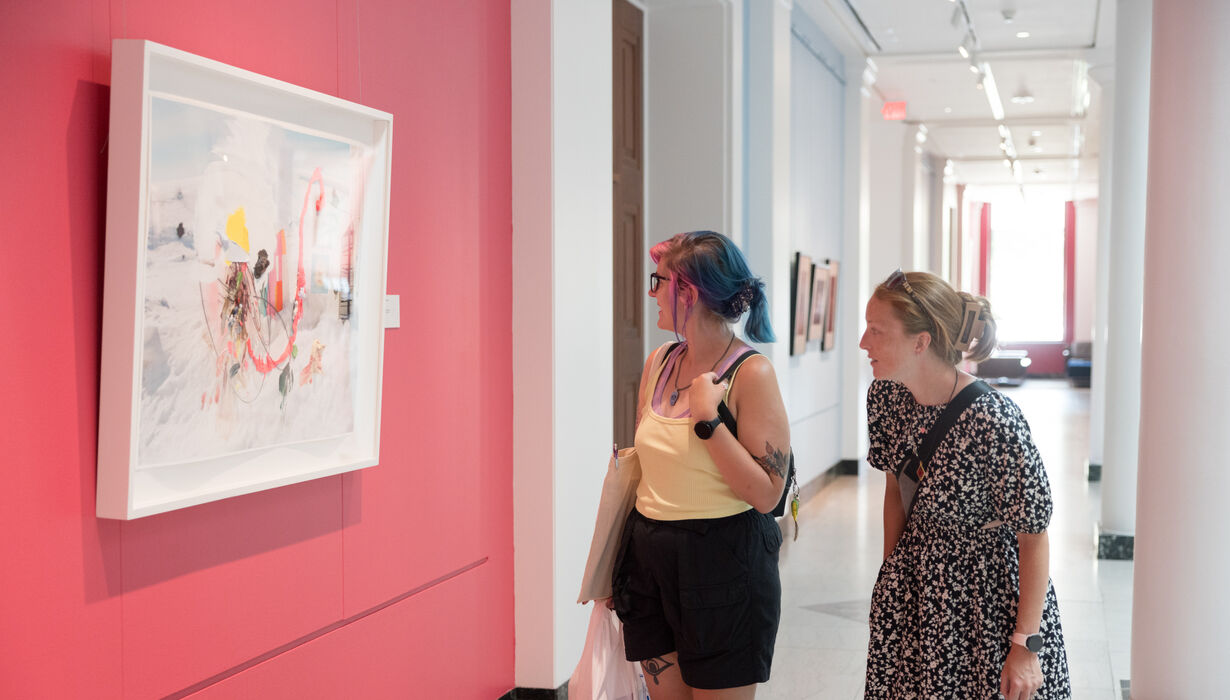 Two young female visitors examine a modern art piece that is hung against a bright pink wall.