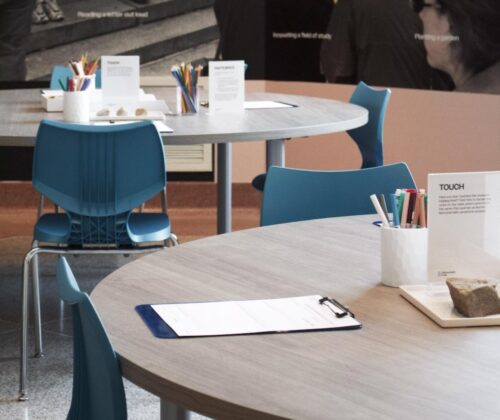 Classroom with round tables and blue chairs. Colored pencils and clipboards rest on top of the desks.