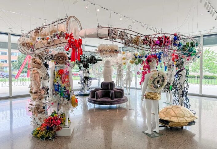 Interior of a modern art exhibition with eclectic sculptures, including detailed wire and mixed media figures adorned with various objects. The space features large windows, a purple sofa set in the center, and is well-lit.