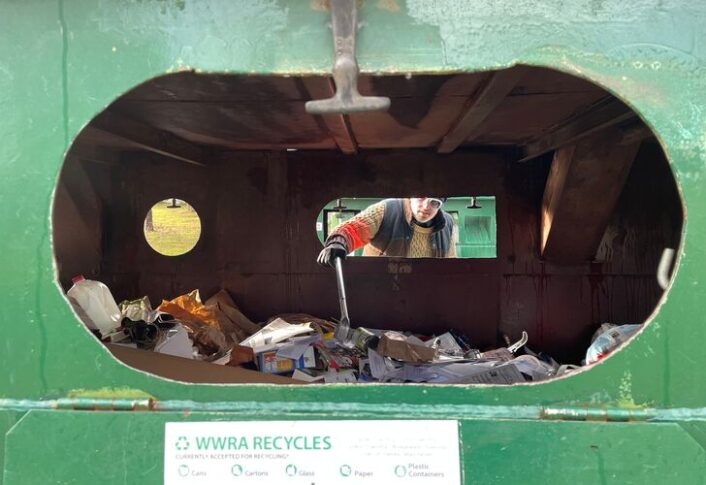 A man is seen through a small window in the center of the image, he is reaching into a large recycling dumpster, using a grabber claw to pick objects out of the trash heap.
