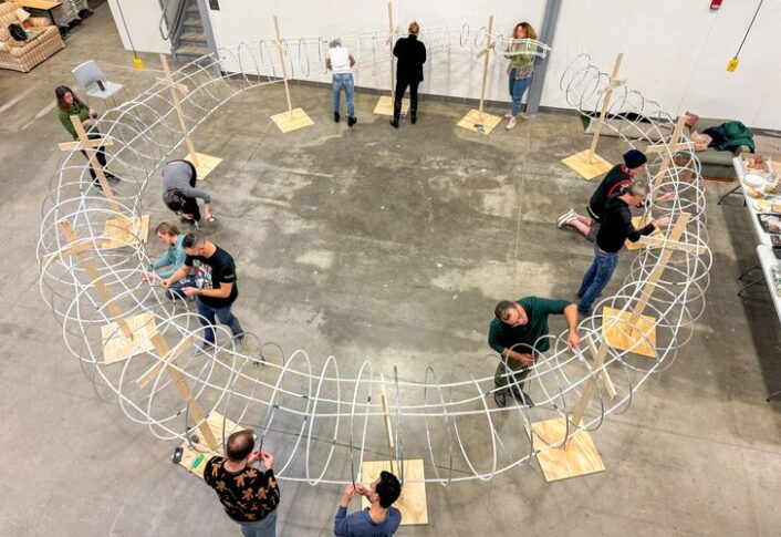 Wide-shot of people helping wrap white coil and wooden stand sculpture