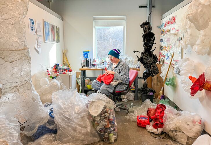 Machine Dazzle sitting in a chair surrounded by garbage working on pieces for his sculpture Ouroboros.