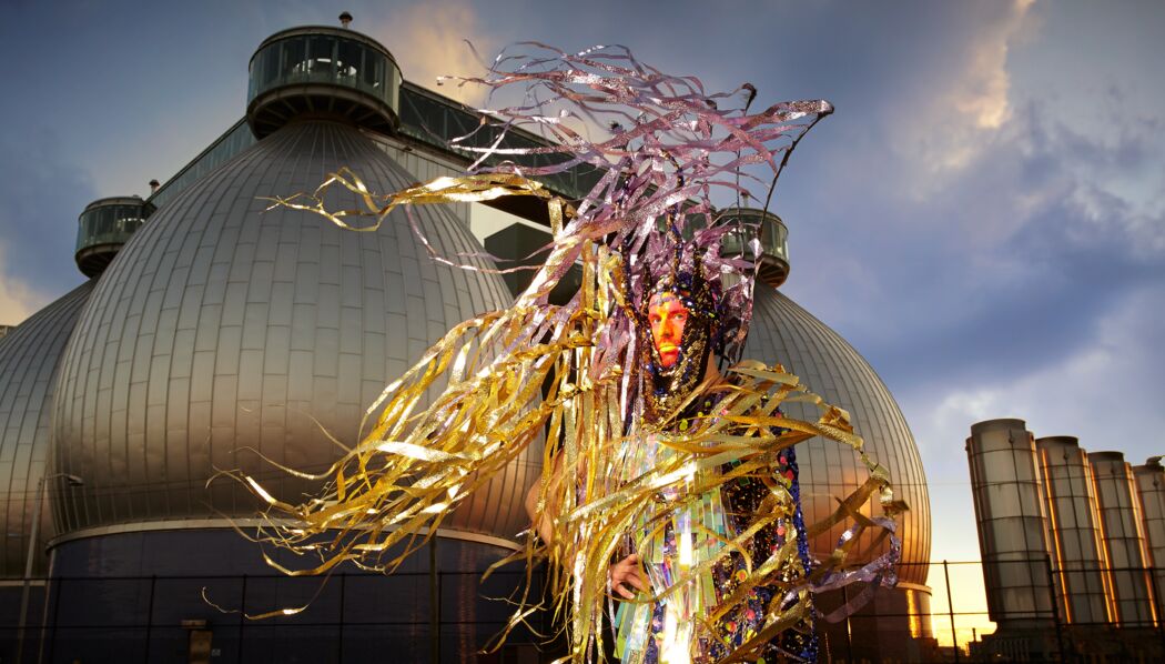 Artist Machine Dazzle dressed in a costume of his design, featuring a lot of yellow and purple metallic fringe blowing in the wind. He stands in front of an industrial, futuristic-looking building that is actually the waste treatment plant in Brooklyn, NY.