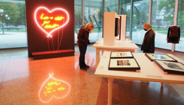 Two people stand in a windowed-room, looking at tables of framed prints and photographs, behind them is a large neon heart on a black wall.