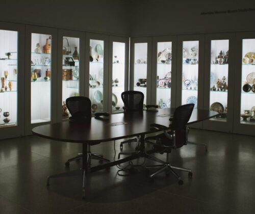 Large table with three chairs in the center of a room with lightbox shelf-lined walls filled with art objects.