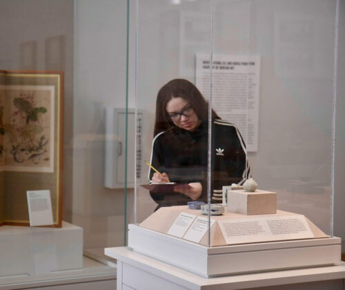 A person stands in front of a small art artifact and takes notes on a clipboard.