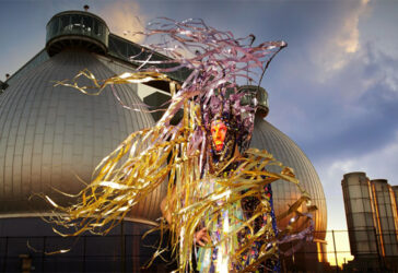 Artist Machine Dazzle dressed in a costume of his design, featuring a lot of yellow and purple metallic fringe blowing in the wind. He stands in front of an industrial, futuristic-looking building that is actually the waste treatment plant in Brooklyn, NY.