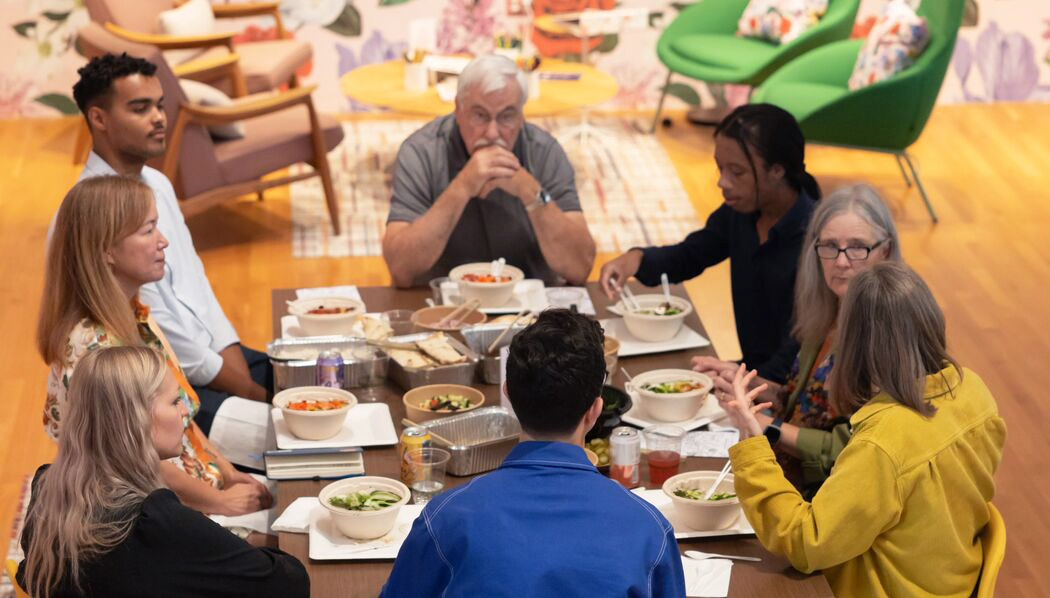 A diverse group of eight people are seated at a rectangular dining table inside a museum gallery space. The table is filled with various dishes and each person is engaged in conversation or eating. A large, colorful floral mural is visible on the wall next to them.