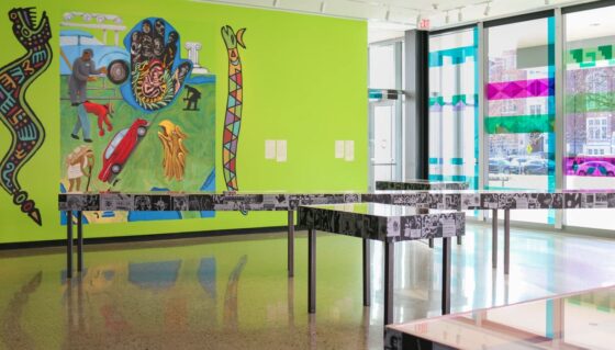 Interior of the LA Raza Art & Medioa Collective with a vibrant, colorful mural on a green wall. Glass doors and windows are reflecting the outside street view. An exhibit display with graphical panels is placed in the foreground.