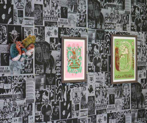 Three framed posters with vibrant designs displayed against a wall covered in various black and white graphic prints. The posters feature bold text and illustrations in pink, green, and yellow tones. A ceramic "witch" placed next to them by Artist Nicole Marroquin.