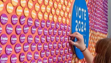 A person placing a circular "I vote" sticker on a colorful board filled with similar stickers, next to a larger blue sign reading "VOTE 2024".