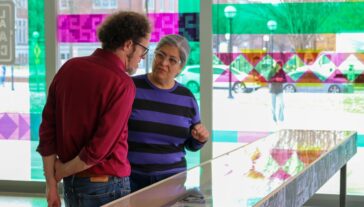 David Choberka and Artist Nicole Maroquin in conversation beside a display case in an art gallery with large, colorful geometric patterns visible through the windows.