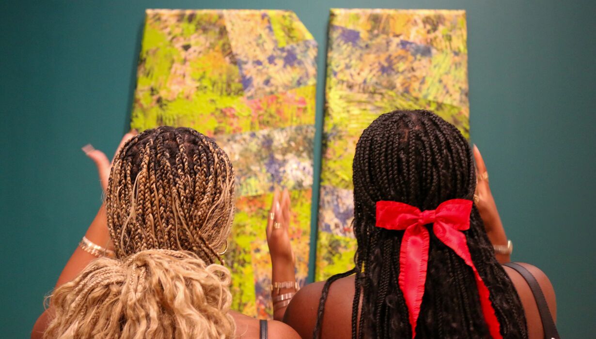 Two people examining abstract paintings in an art gallery, one with braided hair tied with a red ribbon.