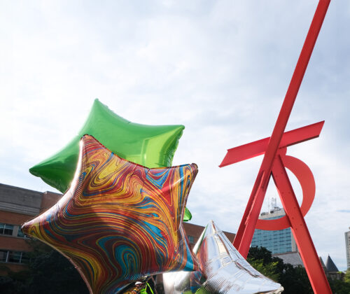 Brightly colored star-shaped balloons float in the foreground against a bright blue sky. A large angular red sculpture stands tall over a green lawn behind them.