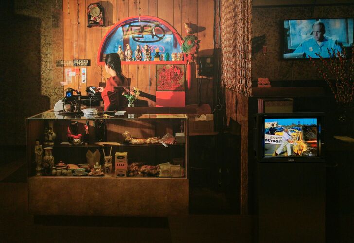 A monitor sits on top of a black platform in front of a wall with a large image of a Chinese restaurant. The monitor displays video footage from 1980s Detroit showing a white man hitting a Toyota with a sledge hammer