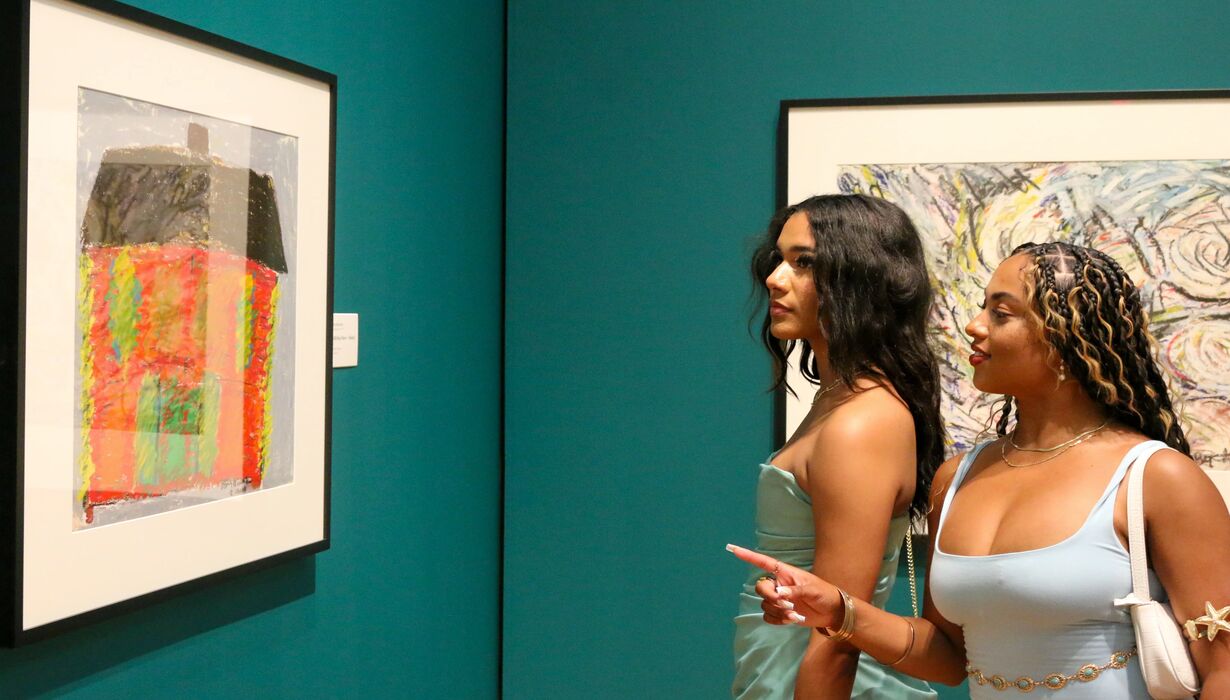 Two individuals viewing artworks in a gallery, with one painting visible on the wall featuring bold, abstract colors. One person is pointing at the piece of art.