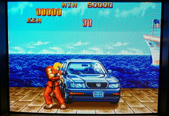 A screen grab from a video game in which a man in an orange karate outfit stands in ready position next to a blue sedan.