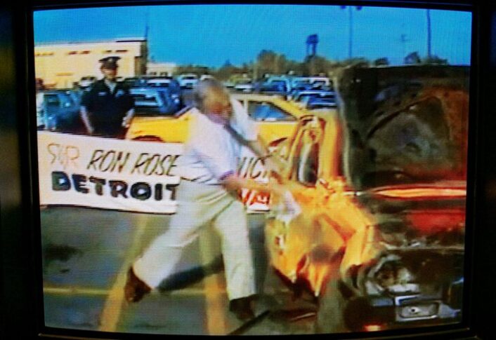 A monitor displaying video footage from 1980s Detroit showing a white man hitting a Toyota with a sledge hammer