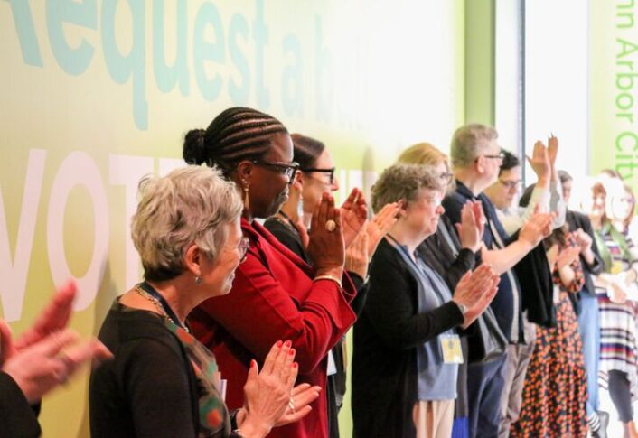 A group of people applauding at an event near a wall with the word "VOTE" displayed prominently.