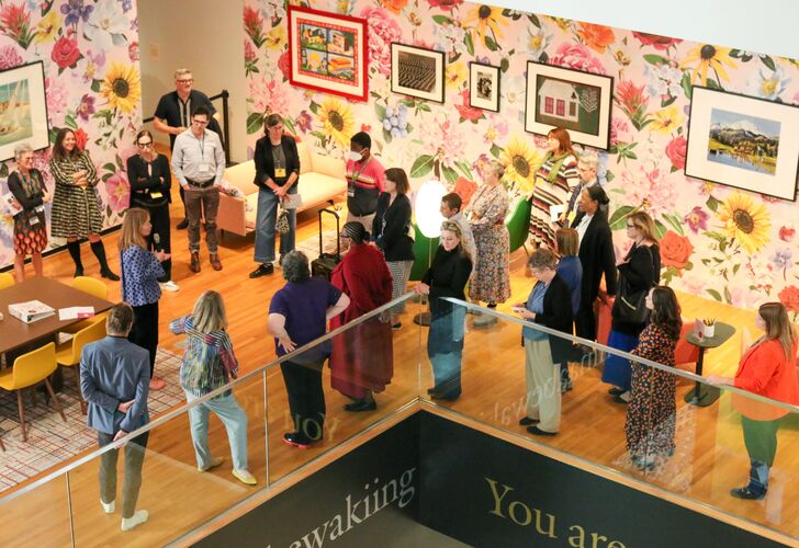 A group of people gathered in a vibrant art exhibition room with floral wallpaper and framed artworks.