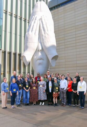 A group of people standing in front of the UMMA entrance beneath a large white sculpture of a face being covered by hands called "Behind Our Walls" by Jaume Plensa.