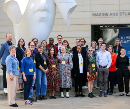 A group of people standing in front of the UMMA entrance beneath a large white sculpture of a face being covered by hands called "Behind Our Walls" by Jaume Plensa.