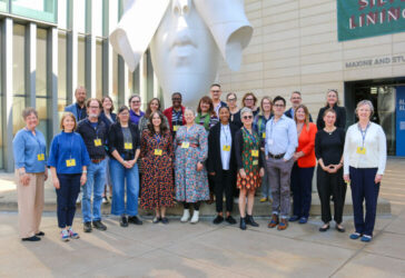 A group of people standing in front of the UMMA entrance beneath a large white sculpture of a face being covered by hands called "Behind Our Walls" by Jaume Plensa.