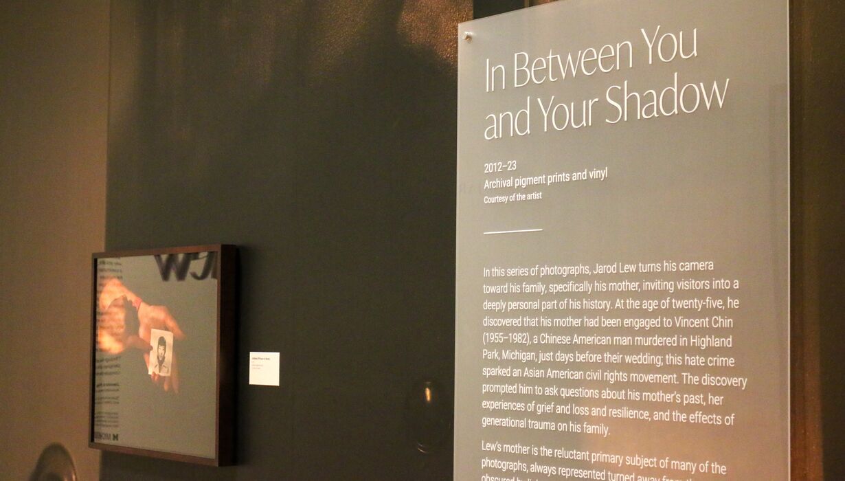 Exhibition panel titled 'In Between You and Your Shadow' displaying text about the artist Jarod Lew's photography and vinyl transfer prints, along with a frame artowrk, in a gallery setting.