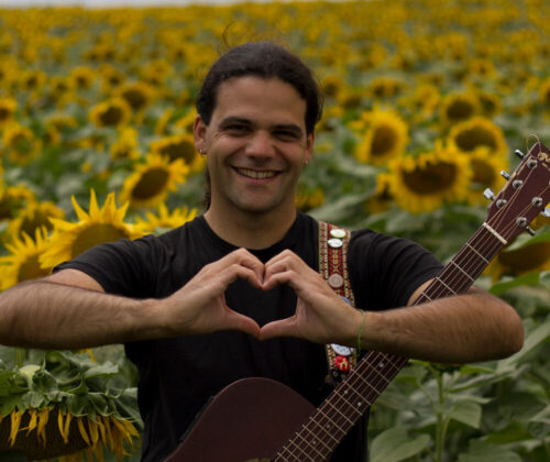 A man with a guitar stands in a field of sunflowers. He is making a heart shape with his hands.