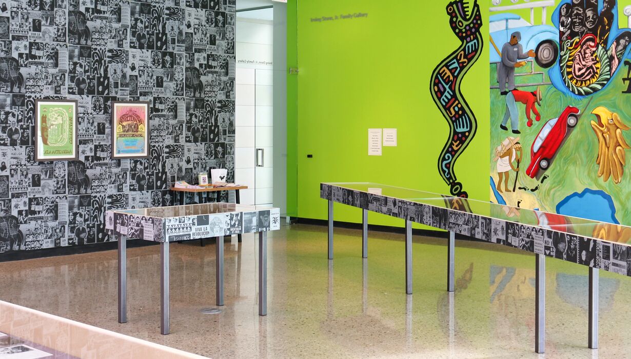 Interior of at he La Raza Art & Media Collective exhibition featuring a vibrant mural and diverse artwork, including a large, colorful wall painting and several smaller framed pieces against a patterned black and white backdrop. Each wall is distinct; one has a bright green color and the other features an extensive wallpaper design. Tables and information placards are present for visitors.