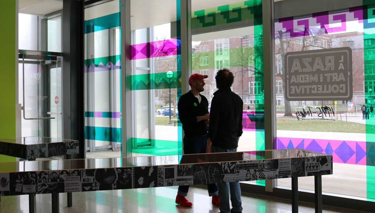 The opening day of the exhibition La Raza showing the custom window artwork with pink, green, and teal colors coming through while two people in silhouette talk in the gallery.