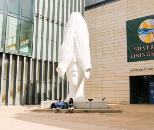 Two people sitting by a large white sculpture of a head in front of the Maxine and Stuart Frankel and the Frankel Family Wing of the University of Michigan Museum of Art, with a banner titled 'Silver Linings' on the building.