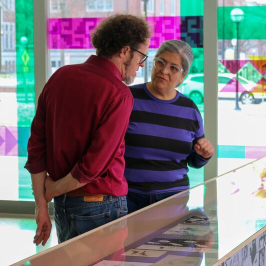 David Choberka and Artist Nicole Maroquin in conversation beside a display case in an art gallery with large, colorful geometric patterns visible through the windows.