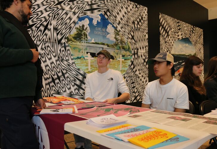 Students gathered outside of the exhibition, sitting at tables with their work displayed.