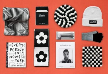 A flatlay of monochrome gift items arranged on a red-orange background. The items include a geometric patterned scarf, a Rhodia notepad, a knit beanie with a patch reading "You Are on Native Land," a floral-patterned scarf, a set of Jotter pens, a pair of black-and-gray gloves, a book titled "Every Person in New York" by Jason Polan, a book titled "God Made My Face" featuring James Baldwin on the cover, and two woven checkerboard trivets.