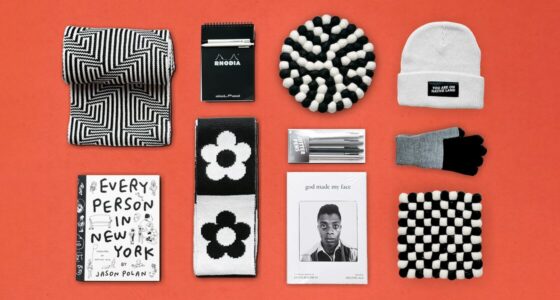 A flatlay of monochrome gift items arranged on a red-orange background. The items include a geometric patterned scarf, a Rhodia notepad, a knit beanie with a patch reading "You Are on Native Land," a floral-patterned scarf, a set of Jotter pens, a pair of black-and-gray gloves, a book titled "Every Person in New York" by Jason Polan, a book titled "God Made My Face" featuring James Baldwin on the cover, and two woven checkerboard trivets.