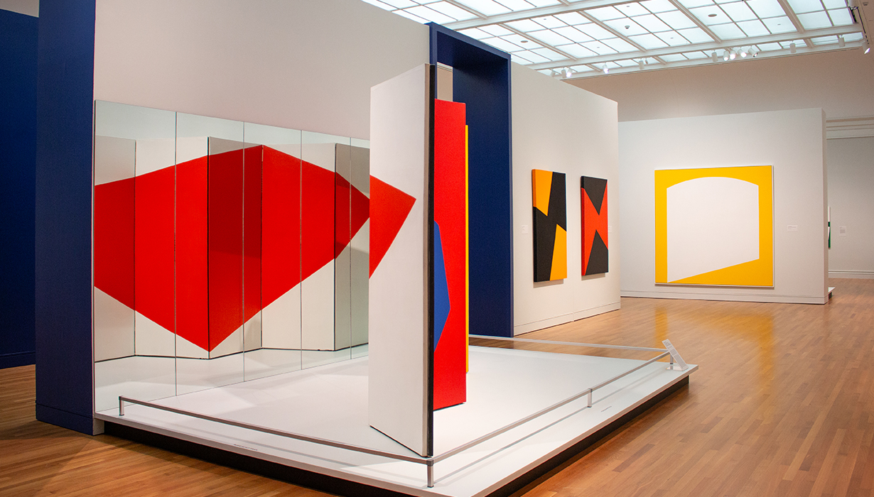 Installation view of a geometric abstract art exhibit featuring red and white panels arranged in a dynamic composition, displayed in a modern gallery setting.