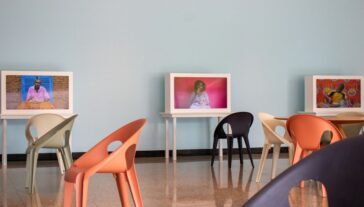 Interior of a room with pastel blue walls featuring three framed digital displays on white tables, each depicting individuals in colorful attire. The room is furnished with modern, multicolored chairs arranged around the tables.