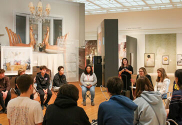 Group of students seated on the floor in chairs in an art gallery at UMMA, attentively listening to a speaker. Surrounded by Jaord Lews works.