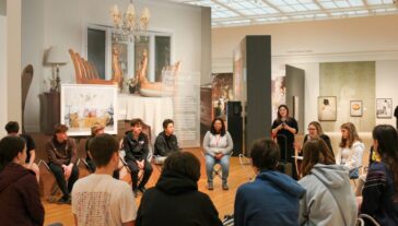 Group of students seated on the floor in chairs in an art gallery at UMMA, attentively listening to a speaker. Surrounded by Jaord Lews works.
